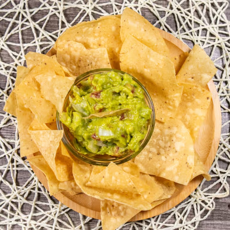 A wooden plate of crispy tortilla chips surrounds a glass bowl of chunky guacamole.
