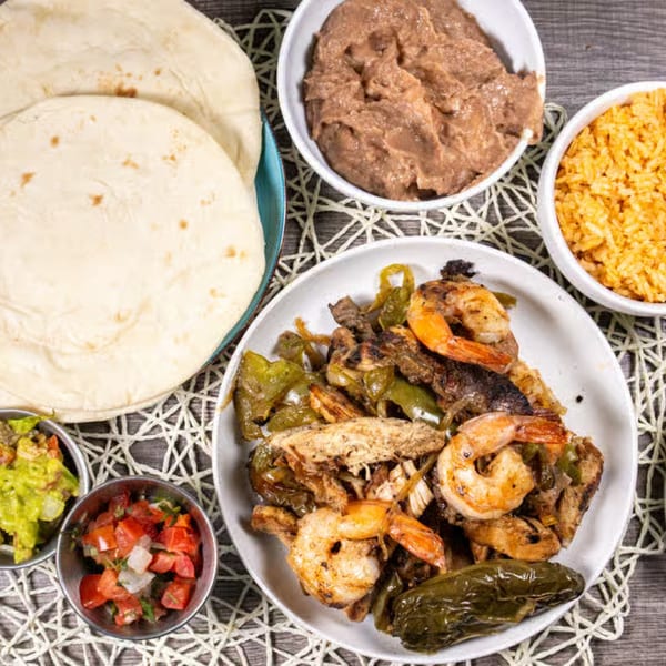 Top-down view of fajitas with shrimp, chicken, and peppers, alongside tortillas, rice, beans, guacamole, and salsa.