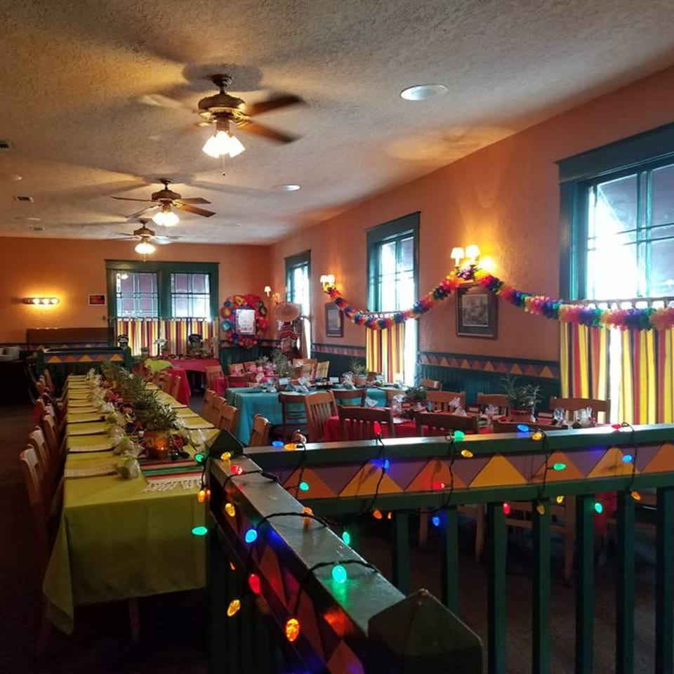 A festive Mexican restaurant dining room with long tables, colorful decorations, and string lights.